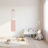 A fabric height chart decal with a scalloped edge design in pink and white, displayed on a wall above a small table with a decorative item on top.
