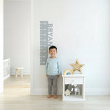 A gray fabric height chart decal with the name 'Bryan' printed on it, adhered to a white wall, next to a coat rack and a wooden stool.